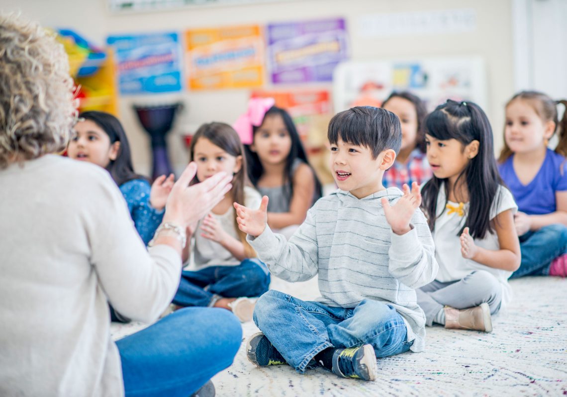 A little boy clapping along with a librarian and other children, all sitting on the floor